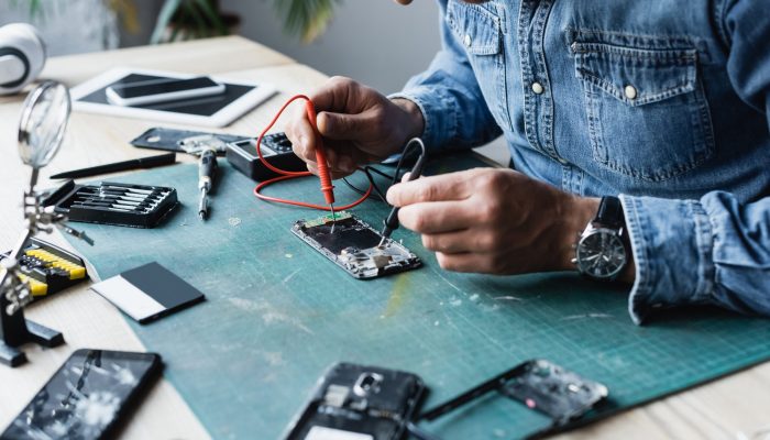 Cropped view of repairman holding sensors of multimeter on disassembled part of mobile phone on